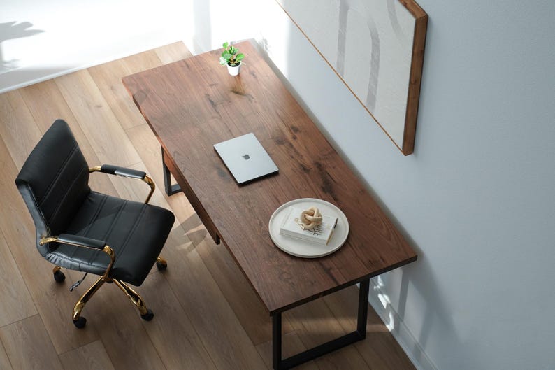 May include: An overhead shot of a home office setup. A dark wood desk with black metal legs holds a laptop, a small plant, and a plate with a decorative object. A black leather office chair with gold accents sits to the left.