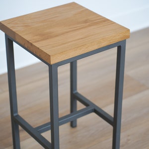 May include: A wooden bar stool with a dark gray metal frame. The stool has a square seat and a simple design.