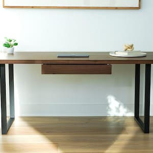 May include: A modern wooden desk with a dark brown finish and black metal legs. The desk features a built-in drawer and a small plant in a white pot. A laptop and decorative items sit on the desk's surface.