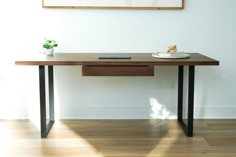 May include: A modern wooden desk with a dark brown finish and black metal legs. The desk features a built-in drawer and a small plant in a white pot. A laptop and decorative items sit on the desk's surface.