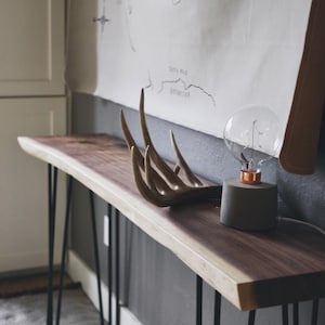 May include: A long, dark wood console table with black hairpin legs. The table has a live edge and is decorated with a set of antlers and a grey concrete lamp with a clear glass bulb.