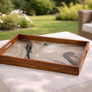 Può includere: Un vassoio rettangolare in legno con cornice marrone scuro e manici integrati. L'interno del vassoio presenta un design astratto beige e nero. Il vassoio è posto su un pouf bianco, con un prato verde e posti a sedere all'aperto sullo sfondo.