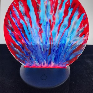 May include: A round, red, blue, and white abstract art piece with a light base. The base has a power switch.