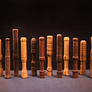 May include: A collection of 14 wooden dowels of varying lengths and diameters, arranged in a row on a black surface. The dowels are made of different types of wood, with some having a natural finish and others having a stained finish.