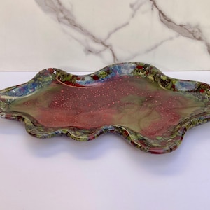 May include: An irregular, wavy-edged glass tray featuring a translucent center with blended shades of deep red and olive green, surrounded by a decorative border of multicolored glass fragments in red, green, blue, and white hues.