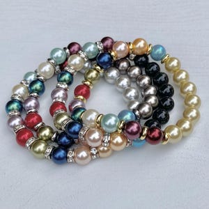 May include: A collection of four stretch bracelets featuring multicolored faux pearls in shades of red, blue, green, gold, silver, and black. Each bracelet is accented with small, clear rhinestone-studded silver or gold-toned metal spacers.