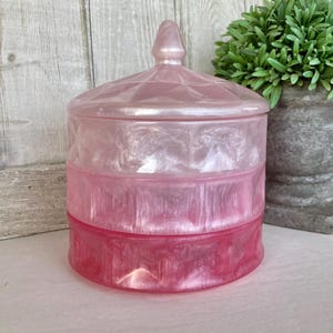 May include: A pink and white iridescent glass jar with a lid. The jar has a layered design with a pink base and a white top.