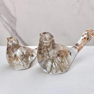 May include: Two clear resin bird figurines, each filled with dried flowers and grasses. The birds have a smooth, glossy finish and are displayed on a white surface. The larger bird has an extended tail.