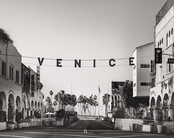 May include: Black and white photograph of Venice, California. The word "VENICE" hangs above the street. Buildings with arches and palm trees line the street. A traffic light and street signs are visible.