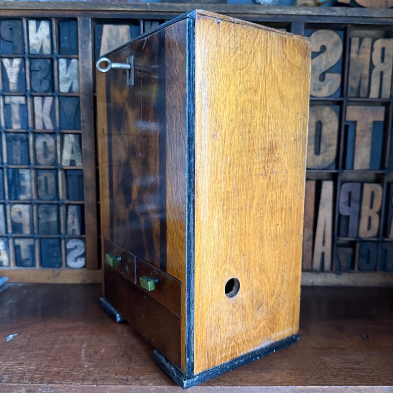May include: A vintage wooden cabinet with two small drawers and a key lock. The cabinet has a light brown finish on the front and sides, with a darker, patterned finish on the left side. The cabinet is set against a backdrop of wooden letterpress blocks.