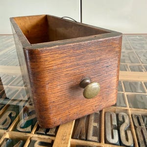 May include: A rectangular wooden drawer with a medium brown wood grain finish and a single rounded brass knob centered on the front panel. The drawer features an open top and a thin metal wire loop attached to the back edge of the frame.