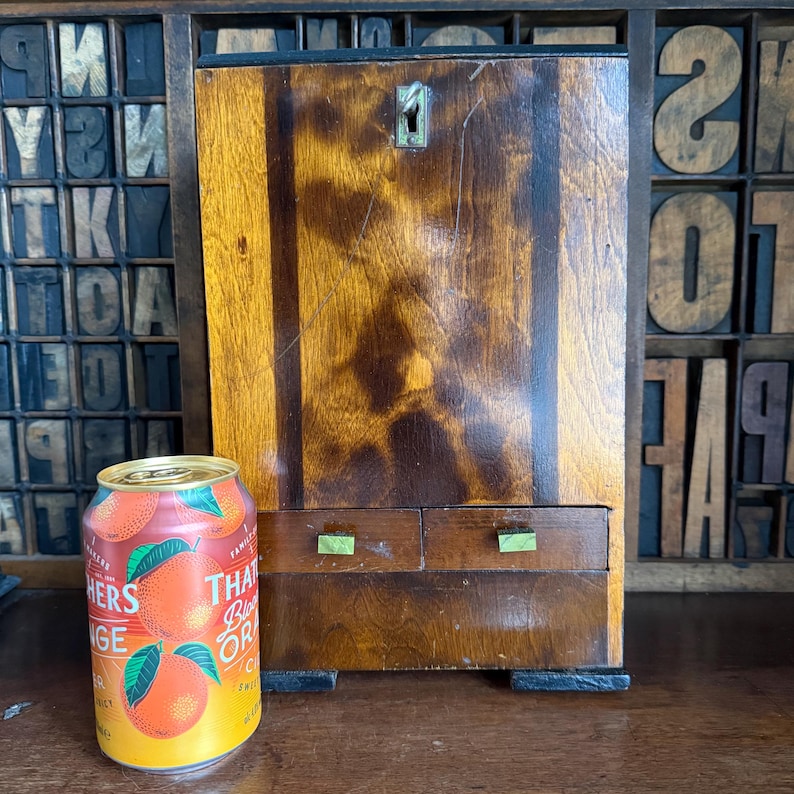 May include: A vintage wooden cabinet with two small drawers and a keyhole. The cabinet has a warm, brown finish with vertical grain patterns. A can of orange soda with the text "THAT BIG ORANGE" is in the foreground.