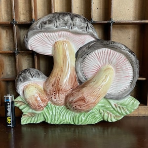 May include: A ceramic wall decor featuring three mushrooms with brown, gray, and pink painted details. The mushrooms are depicted with stems and caps, and are set against a green leafy background.