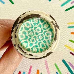 May include: A round cabinet knob featuring a distressed off-white metal frame with visible dark brown paint chips. The center is inlaid with a smooth, glossy glass dome displaying a repeating teal and white floral pattern across the surface.