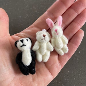 May include: Three small plush toys: a black and white panda, a white teddy bear, and a white bunny with pink ears. The toys are sitting on a person's hand.