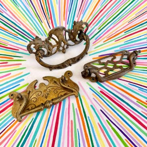 May include: Three antique brass drawer pulls with distinct designs. One features ornate scrollwork, another has a geometric lattice pattern, and the third displays an engraved floral motif. Each piece includes a swinging handle and mounting holes.