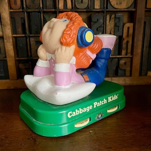 May include: A green Cabbage Patch Kids music box featuring a red-haired doll wearing blue jeans and a pink shirt. The doll is lying on a white pillow and wearing headphones. The music box has a dial to adjust the volume.