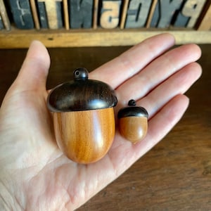 May include: Two wooden acorn-shaped ornaments, one larger than the other. The larger acorn is made of light brown wood with a dark brown top. The smaller acorn is made of light brown wood with a dark brown top.