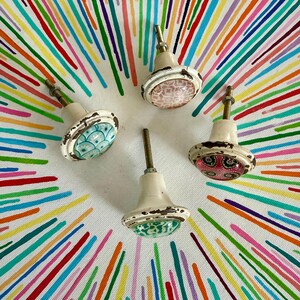 May include: Four round cabinet knobs with white bases and distressed metal hardware. Each knob features a unique circular top with distinct patterns: one with blue scales, one with pink geometric shapes, one with green floral motifs, and one with pink floral designs.