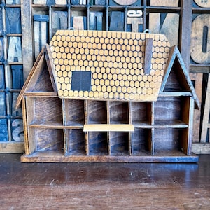 May include: A wooden house-shaped display shelf featuring multiple cubby compartments. The roof section has a decorative honeycomb pattern, a small rectangular black window, and a vertical wooden chimney piece attached to the front surface.