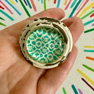 May include: A circular knob featuring a distressed white and brown outer rim with a smooth, glossy center insert. The center displays a repeating teal and white geometric pattern under a clear, slightly domed glass surface.