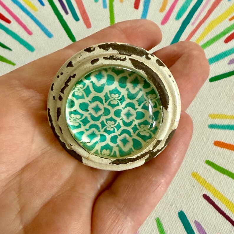 May include: A circular knob featuring a distressed white and brown outer rim with a smooth, glossy center insert. The center displays a repeating teal and white geometric pattern under a clear, slightly domed glass surface.