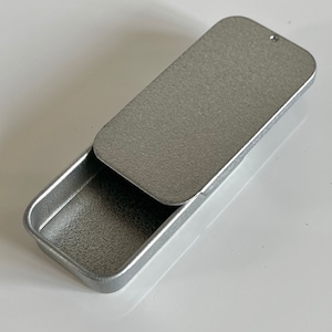 May include: A silver metal tin with a hinged lid. The lid is open and the tin is empty.