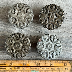 May include: Four antique metal buttons with intricate floral patterns. The circular buttons feature a central flower motif encircled by swirling leaf designs. The buttons are in silver and bronze tones, with a vintage appearance.