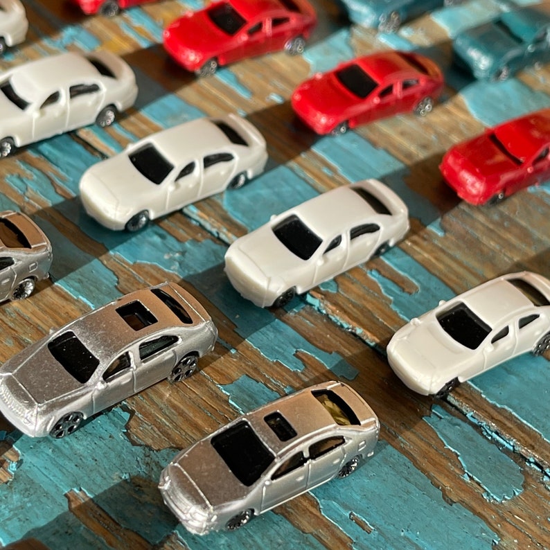 May include: A collection of miniature toy cars in various colors, including silver, white, red, and blue. The cars are arranged on a wooden surface with a blue and brown pattern.