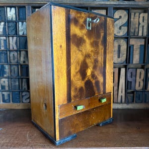May include: An antique wooden cabinet with a keyhole and two small drawers. The cabinet has a dark wood finish with lighter wood panels and green drawer pulls. The cabinet is rectangular and sits on a wooden surface.