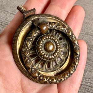 Könnte beinhalten: Antiker Schubladengriff aus Messing mit kreisförmigem Design. Der Griff hat ein zentrales Blumenmotiv, umgeben von einem dekorativen Rand und einer Schraube zur Befestigung. Das Messing hat eine verwitterte Patina.