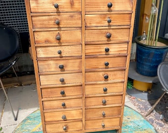 Könnte beinhalten: Ein Holzschrank mit 18 Schubladen, jede mit einem schwarzen Knopf. Der Schrank ist aus hellem braunem Holz und hat einen rustikalen Look.