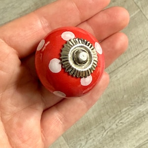 May include: A red ceramic cabinet knob with white polka dots and a silver metal base. The knob is round and has a glossy finish, suitable for adding a decorative touch to furniture.