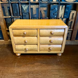 May include: A small, wooden cabinet with six drawers, each with a small, round knob. The cabinet is made of light-colored wood and has a smooth, varnished finish. The cabinet sits on four small, rounded feet.