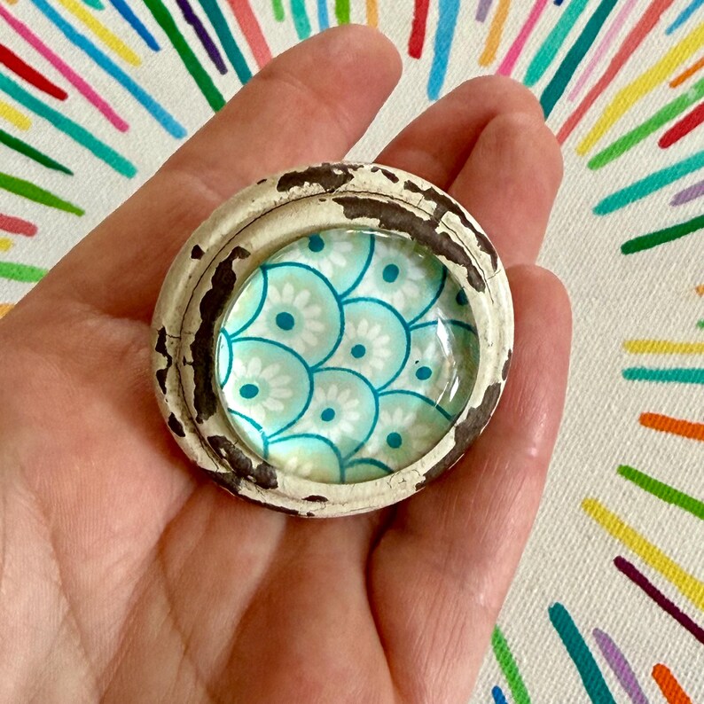 May include: A circular knob featuring a distressed white and dark brown painted wooden frame surrounding a glass insert. The insert displays a repeating pattern of overlapping teal and white scalloped circles with small floral centers.