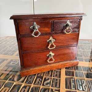 May include: A small, antique wooden chest of drawers with four drawers. The chest is a rich, dark brown color with brass-toned hardware. The drawers have decorative pulls. The chest is sitting on a surface with letterpress printing blocks.