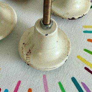 May include: A set of off-white cabinet knobs with a distressed, crackled finish and visible brown rust spots. Each knob features a central threaded metal bolt with a hexagonal nut attached, showing signs of wear and age on the surface.
