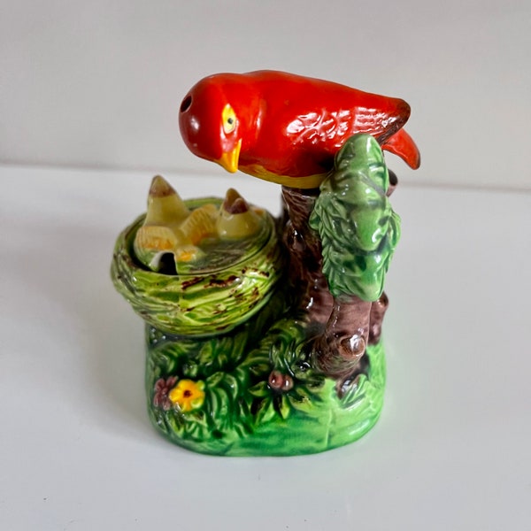 Puede incluir: Figura de pájaro de cerámica con cuerpo rojo, pico amarillo y un nido con pajaritos. La figura está posada en un tronco de árbol con follaje y hierba verdes. La base es verde con una flor amarilla.