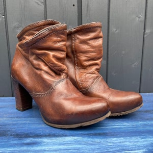 May include: A pair of brown leather ankle boots with a rounded toe and a stacked block heel. The boots have a scrunched design around the ankle and a sturdy sole. The boots are approximately 6 inches tall.
