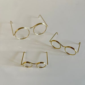 May include: Three pairs of gold wire-framed spectacles of different sizes. The spectacles are arranged in a triangular pattern on a white surface.