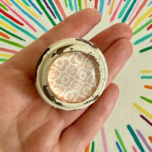 May include: A round cabinet knob featuring a distressed off-white painted metal frame with visible dark brown chips. The center is a smooth, circular glass insert displaying a repeating light pink and white quatrefoil geometric pattern.