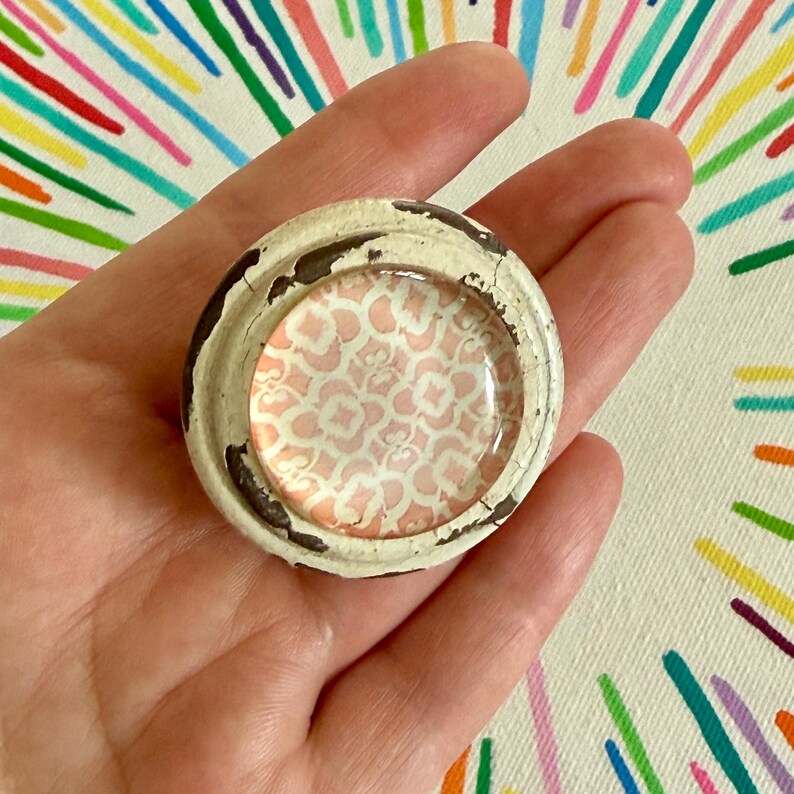 May include: A round cabinet knob featuring a distressed off-white painted metal frame with visible dark brown chips. The center is a smooth, circular glass insert displaying a repeating light pink and white quatrefoil geometric pattern.