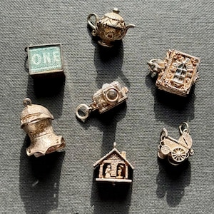 Könnte beinhalten: Eine Sammlung von sieben silberfarbenen Metallanhängern, darunter eine Teekanne, ein quadratischer Anhänger mit dem Wort ONE, eine Kamera, ein detailliertes Buch, ein Ritterhelm, ein kleines Haus mit Figuren und ein Kinderwagen im Vintage-Stil.