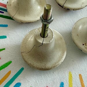 May include: A set of off-white ceramic knobs with a rounded, flared shape. Each knob features a central threaded metal bolt with a hexagonal nut attached. The ceramic surfaces show visible fine cracks and dark, weathered edges throughout the finish.