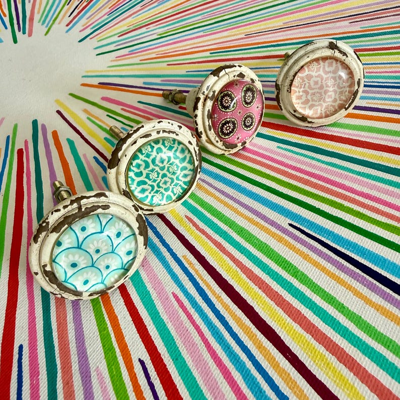May include: Four round cabinet knobs with distressed white metal frames and glass inserts. Each knob features a unique colorful pattern: one with blue and white scales, one with green floral motifs, one with pink and black circles, and one with a light pink geometric design.