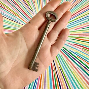 May include: An antique metal key with the word "RELIANCE" and "KEY CO. NY" engraved on the circular head. The key is long and slender, with a detailed key bit. The key is held against a colorful striped background.
