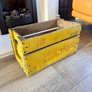 May include: A rectangular wooden crate painted in a weathered yellow finish. The side features the red text "MEUSE" and the number "66" printed in a faded font. The crate has visible wood grain, metal nail heads, and open slats on the sides.