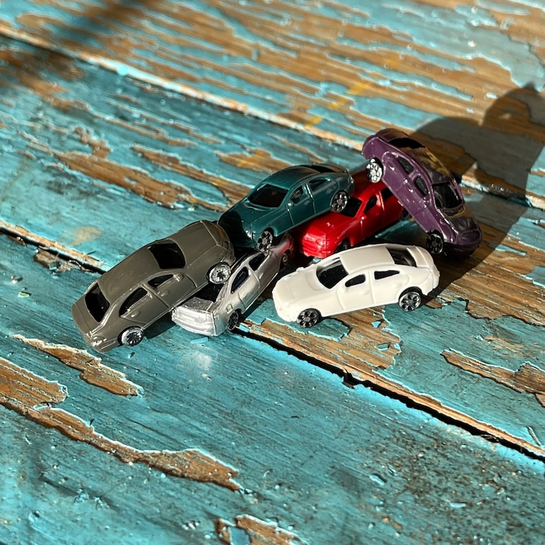 May include: A collection of five miniature toy cars in various colors, including gray, white, blue, red, and purple. The cars are arranged in a pile, with some overlapping. The cars are made of plastic and have realistic details.