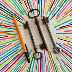 May include: An orange pen and three antique keys are arranged on a colorful background. The keys are various sizes and shapes, with visible wear and tear. The background features a pattern of radiating, multicolored lines.