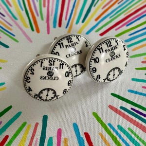 May include: Three white ceramic cabinet knobs with a clock face design. Each knob features black numerals, hands, and the text "PARIS CLOCKS." The knobs are arranged on a white surface with a colorful, abstract background.